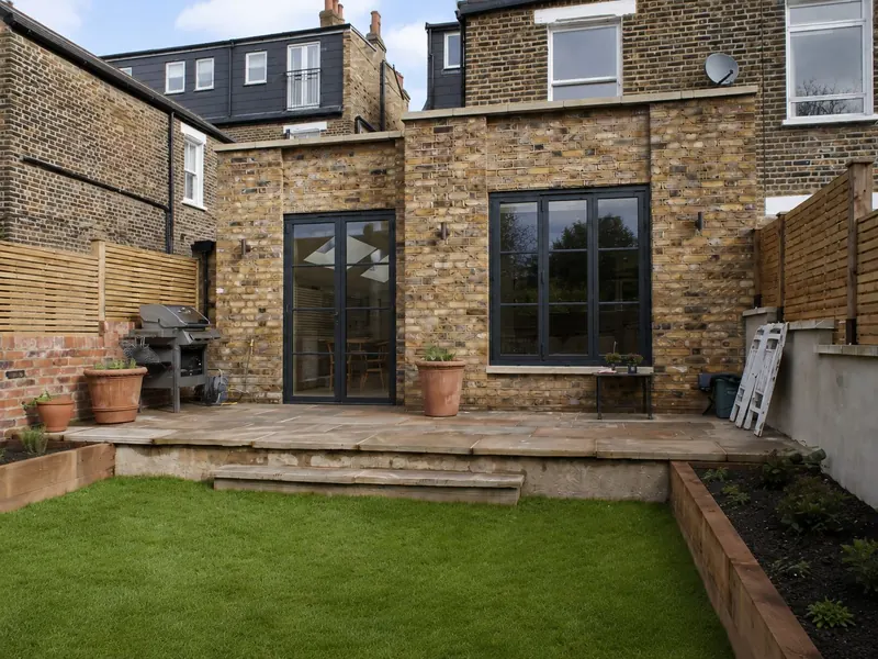 A London stock brick rear extension with black steel-framed doors and a sandstone terrace